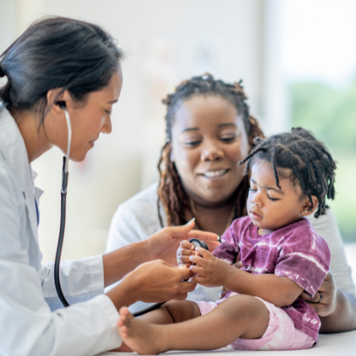 Doctor using a stethoscope to examine a young child sitting on a caregiver’s lap, with both adults focused on the child in a clinical setting.